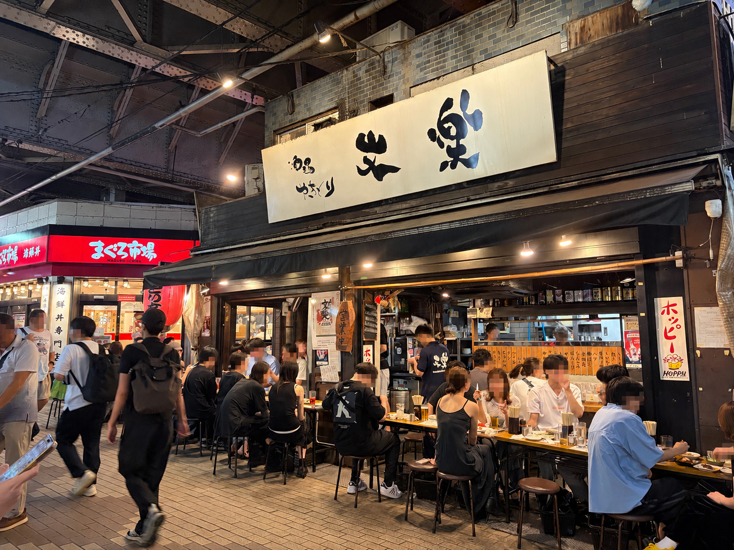 夜の繁華街にある居酒屋の外観。大きな日本語看板の下にカウンター席とテーブル席が並び、多くの客が食事をしている。