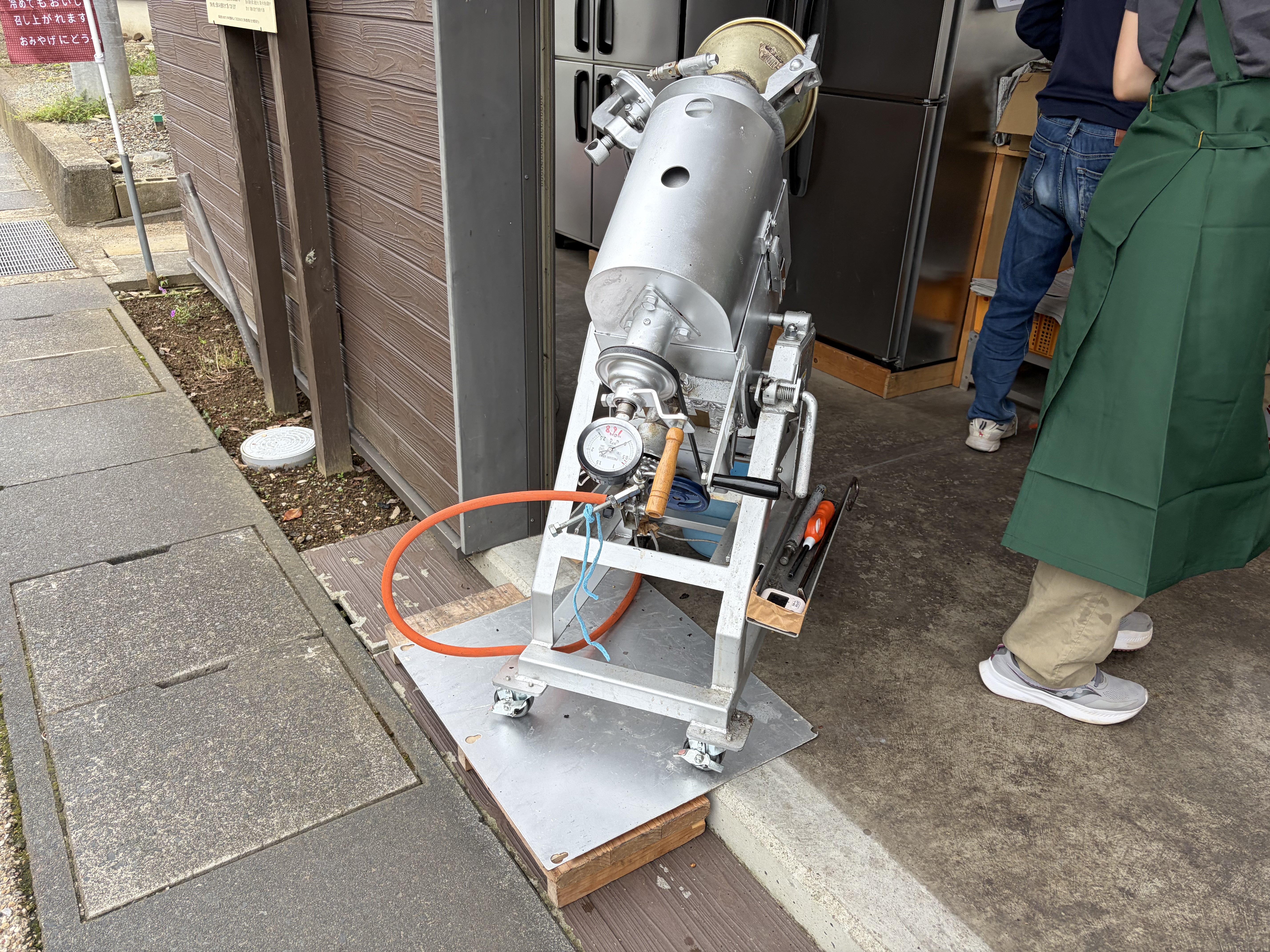建物の出入口前に、金属製の機械とホースが置かれ、そばに二人の人物の下半身が写っている。