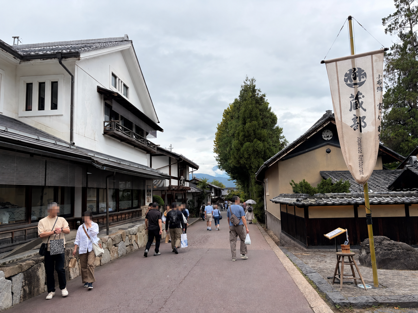 白壁の建物や瓦屋根の家並みが続く通りを、数人の歩行者が歩いている。通り沿いには木製の案内旗や植栽が見える。