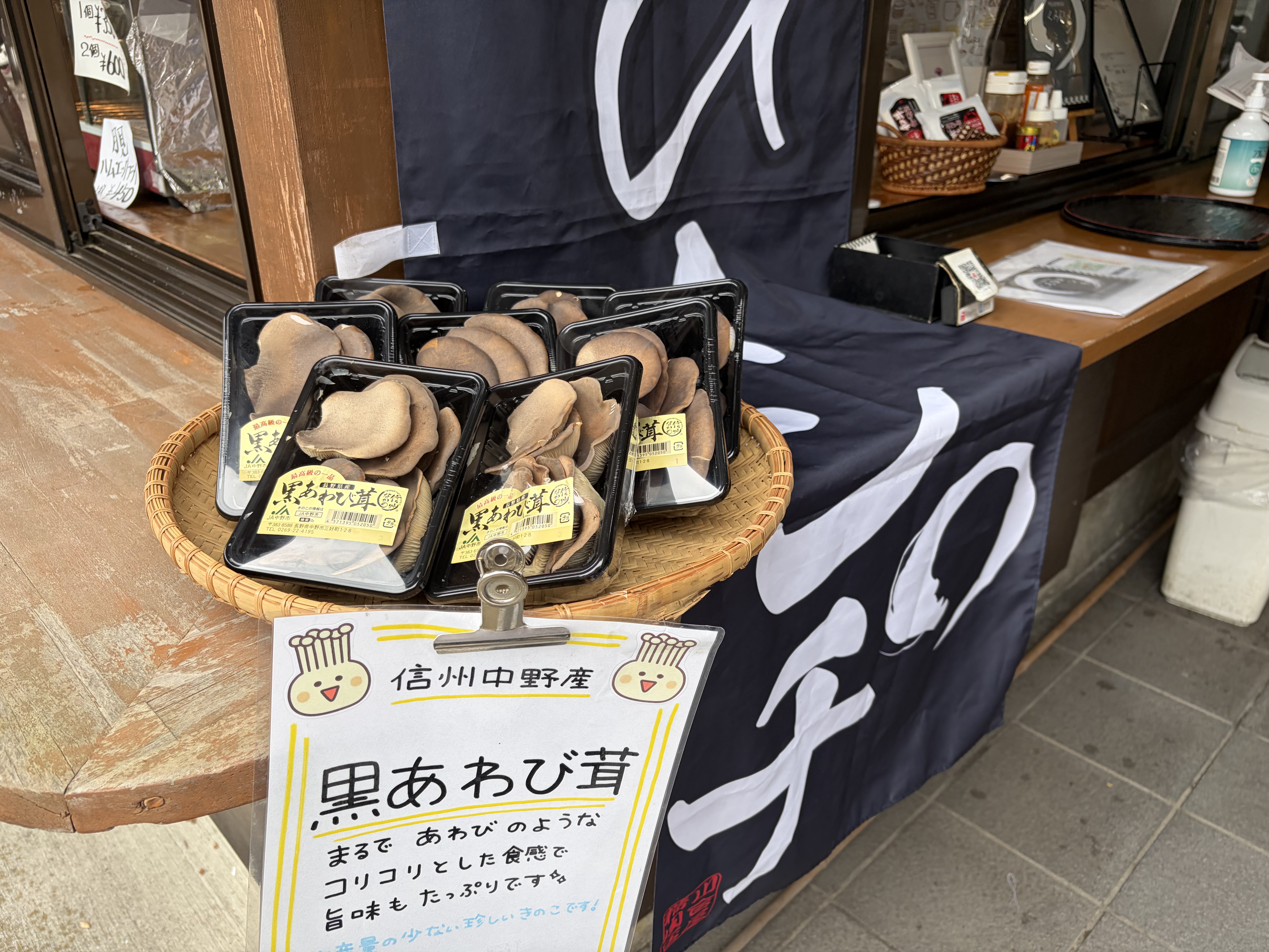 店先に、黒あわび貝と書かれた食品のパックがかごや台の上に並び、筆文字ののれんと手書きの案内板が見える。