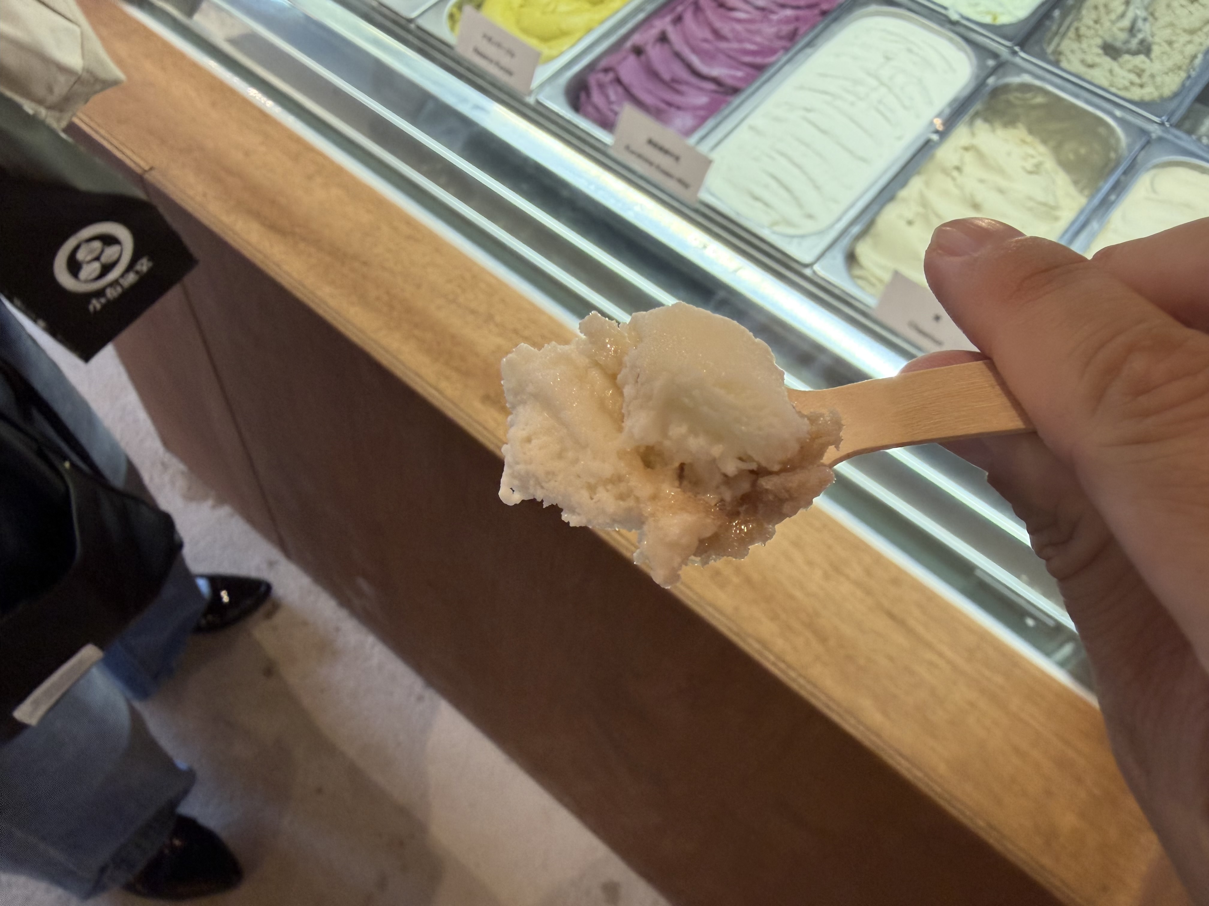 木製スプーンですくわれたジェラートの一口分と、ショーケースに並ぶ複数のフレーバー。