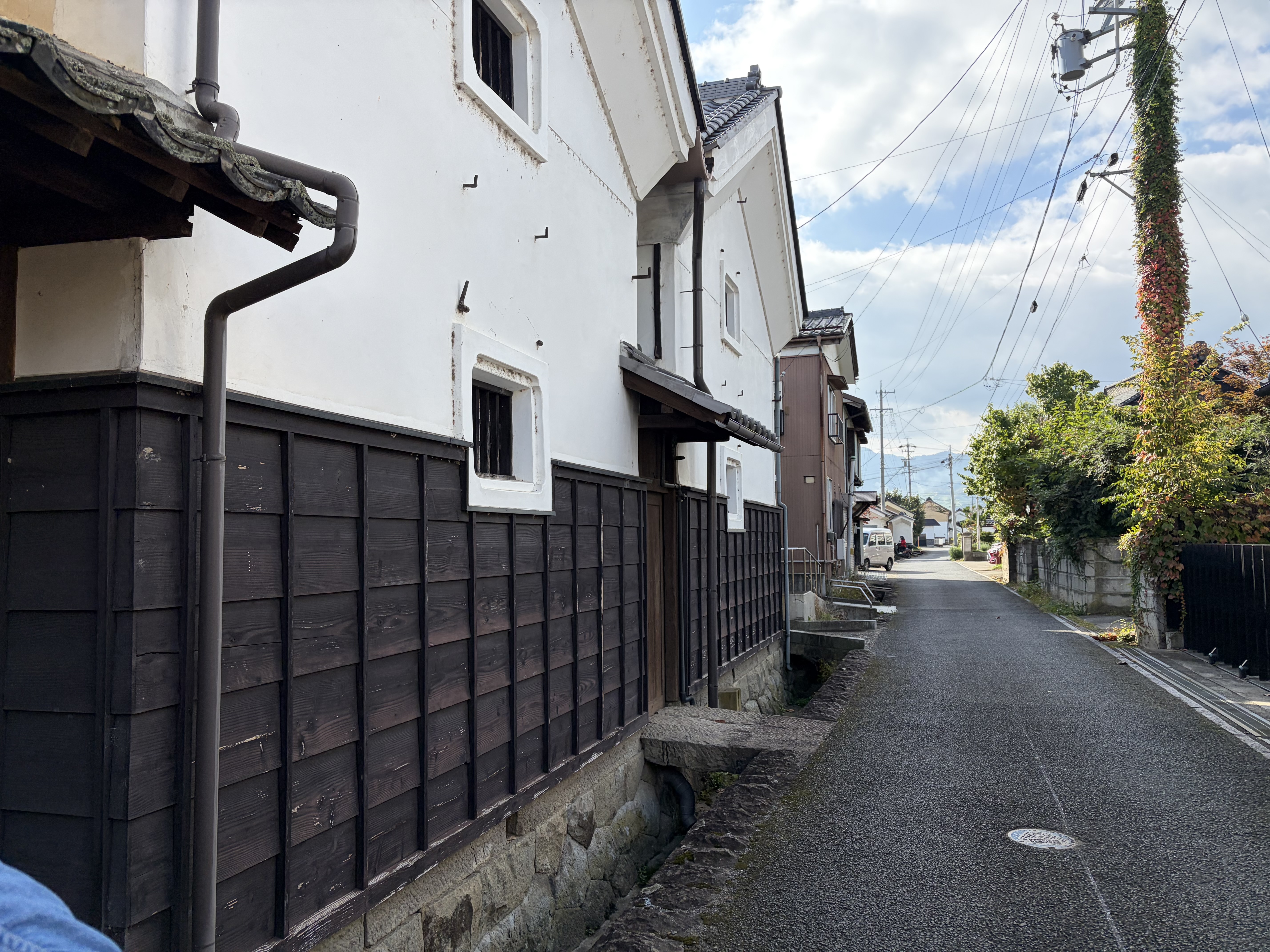 白壁と黒い木製建具の建物が並ぶ、電線のある細い舗装路。青空の下、道の先に住宅や木々が見える。