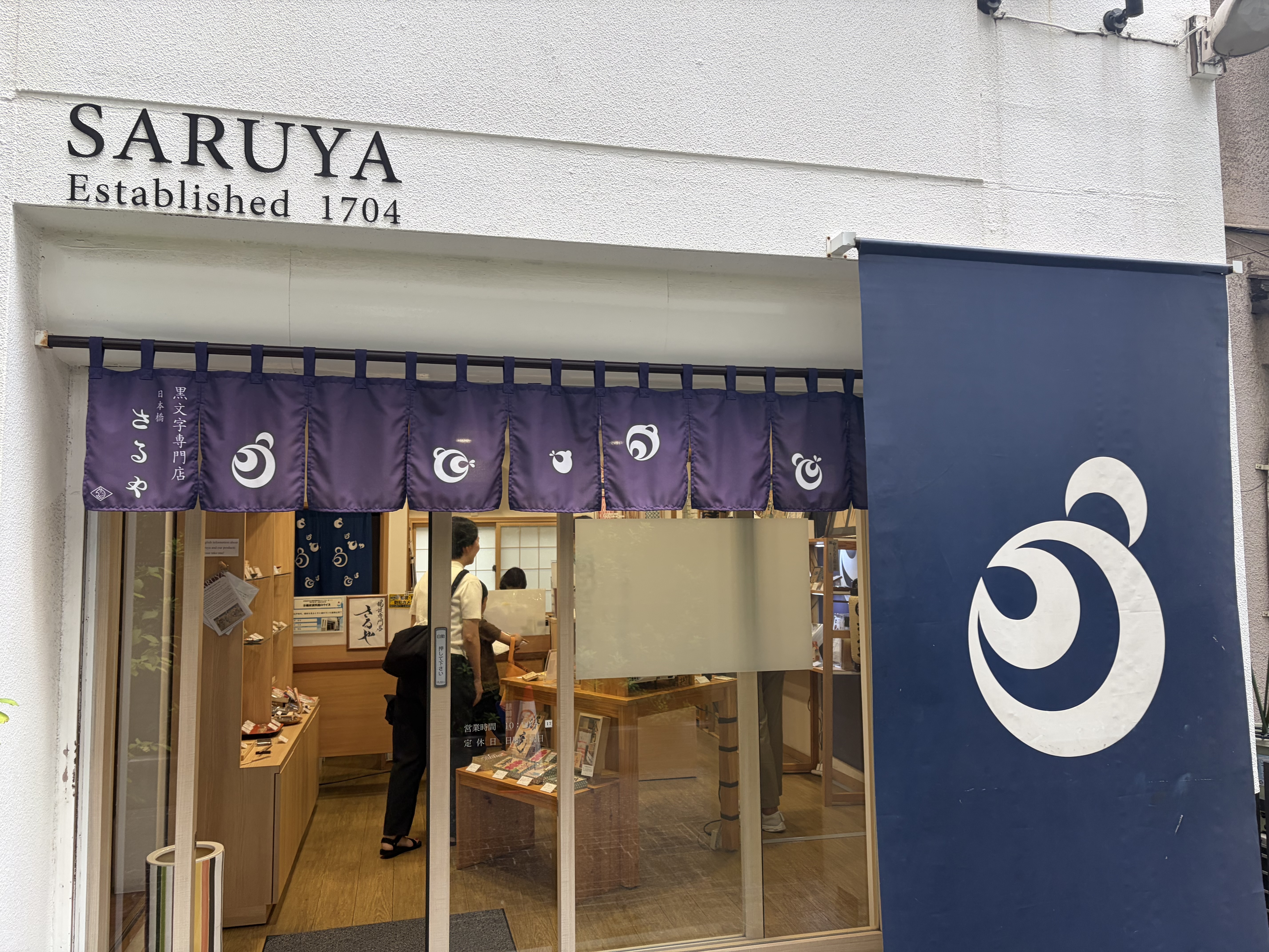 「SARUIYA Established 1704」と書かれた店舗の外観。紫の暖簾と白いロゴが掲げられ、ガラス越しに店内の商品棚が見える。