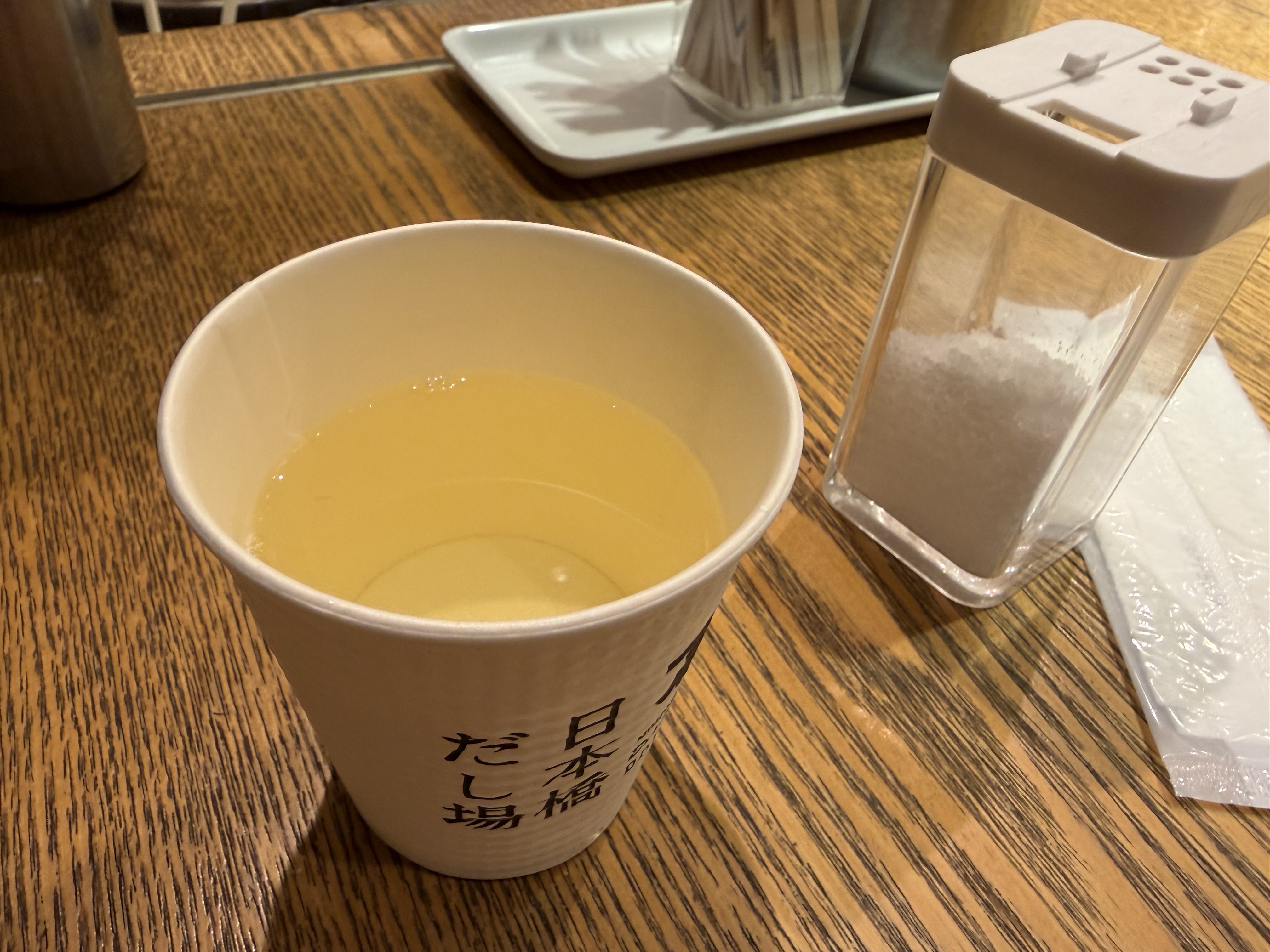 木目のテーブルの上に置かれた、黄色みのある飲み物入りの紙カップ。