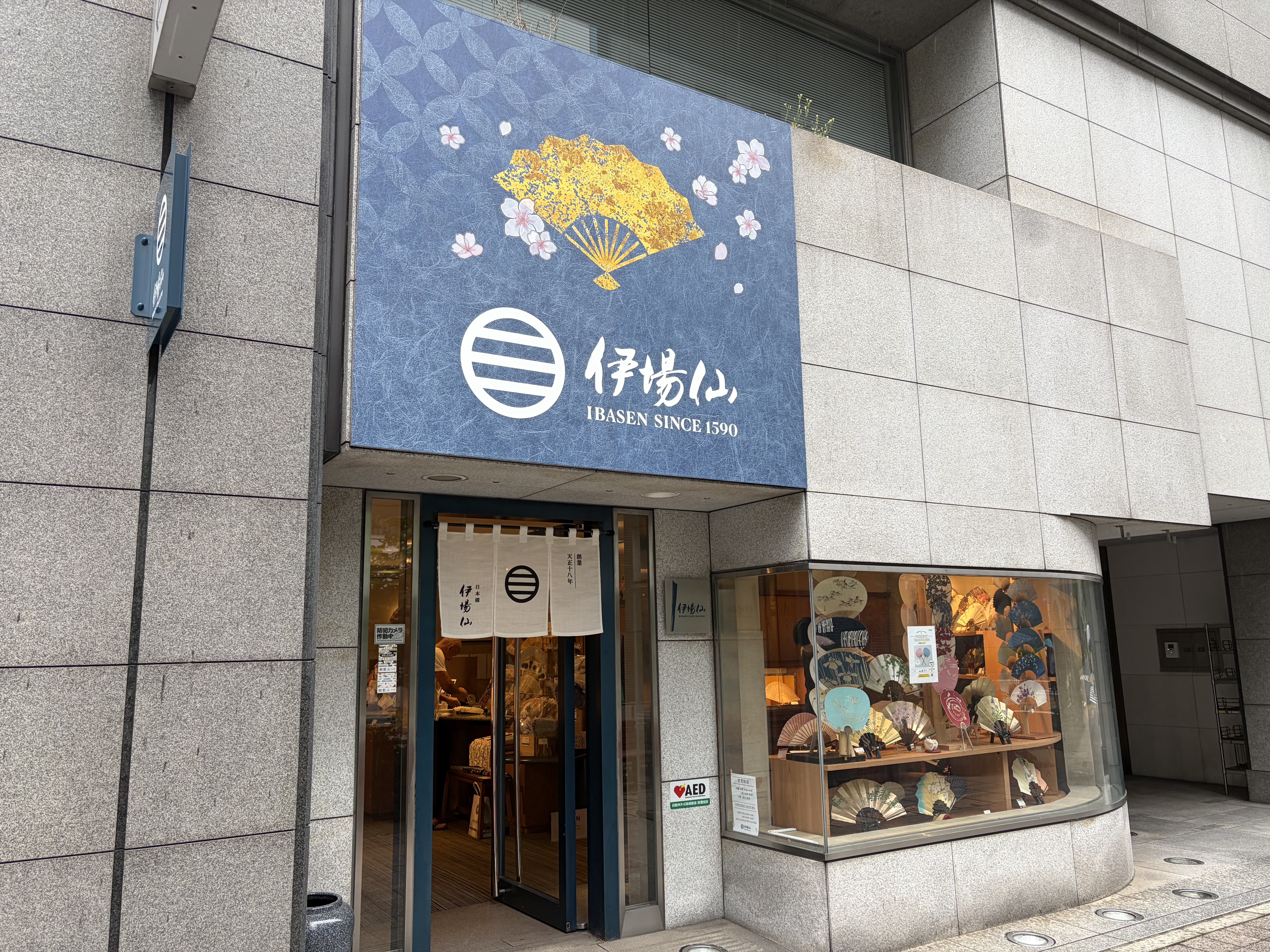 石張りの建物にある和風店舗の外観。青い看板に扇と花の絵が描かれている。