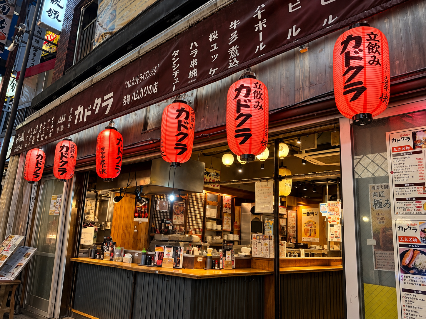 赤い提灯が並ぶ大衆酒場「カドクラ」の店頭。開いた店舗からカウンターとメニューが見えている。