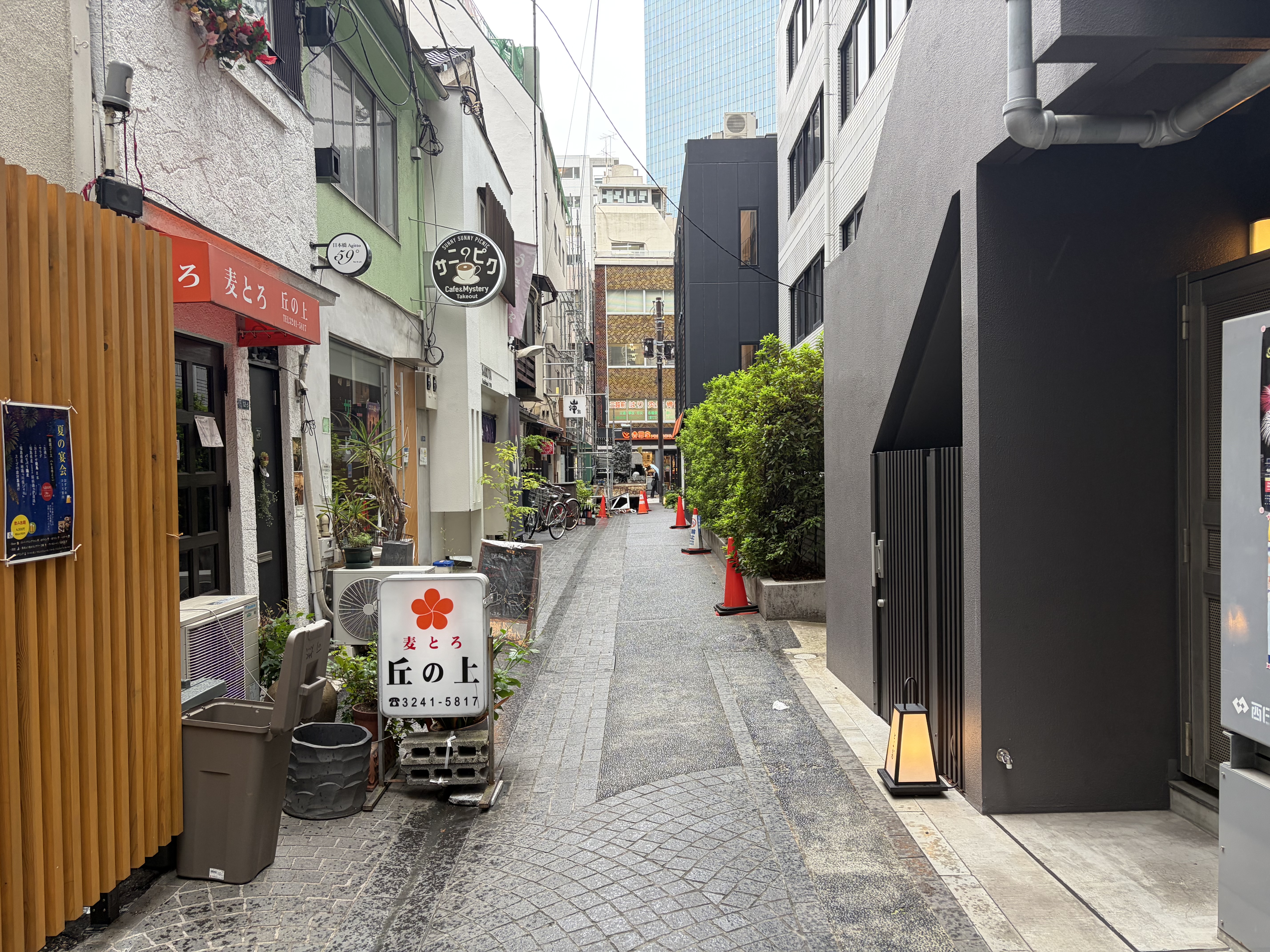 飲食店の看板や植木、自転車が並ぶ日本の細い路地。奥に高層建築が見える。
