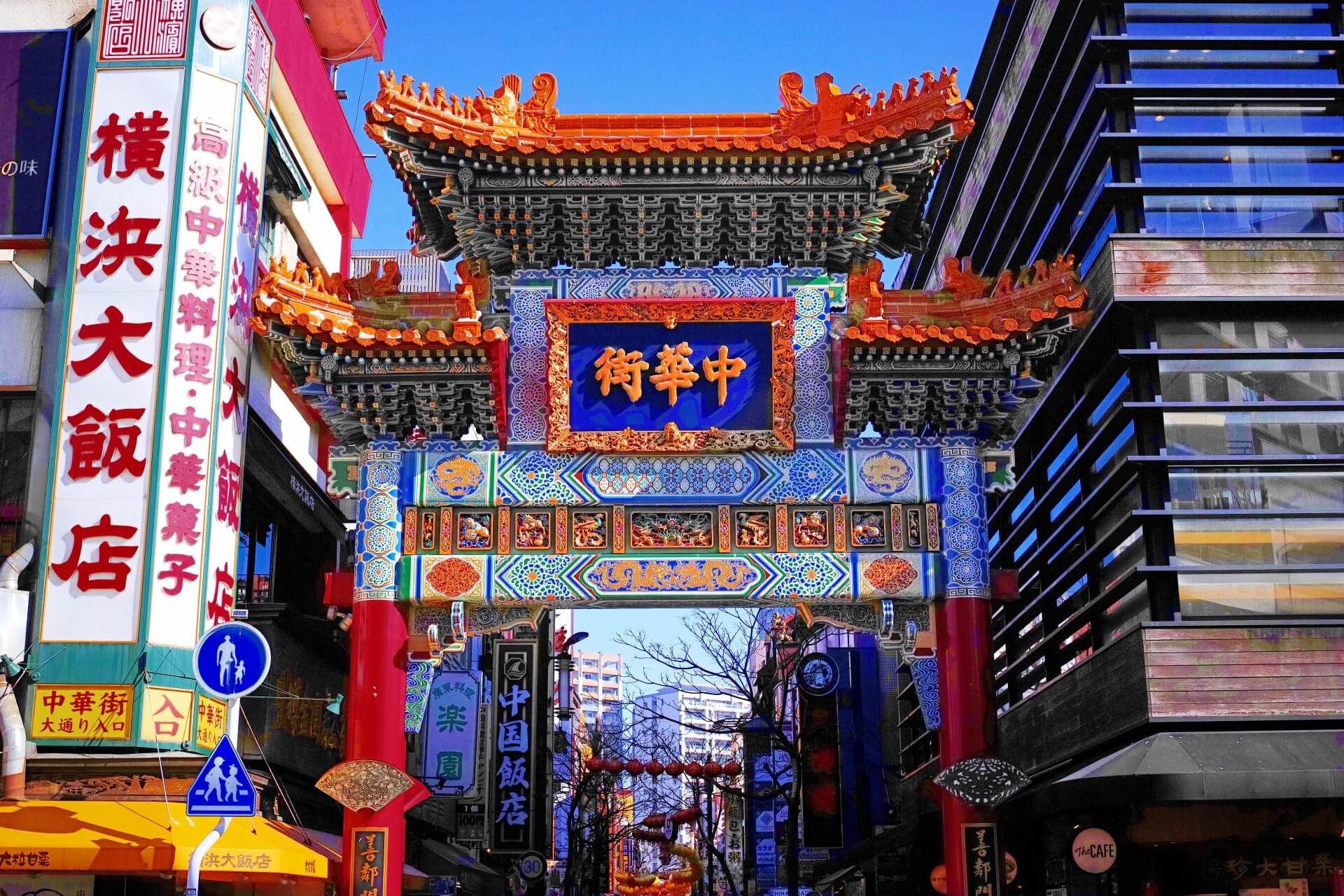 色鮮やかな装飾と漢字の扁額を備えた中国風の門が、商店や現代的な建物に囲まれた通りに立っている。