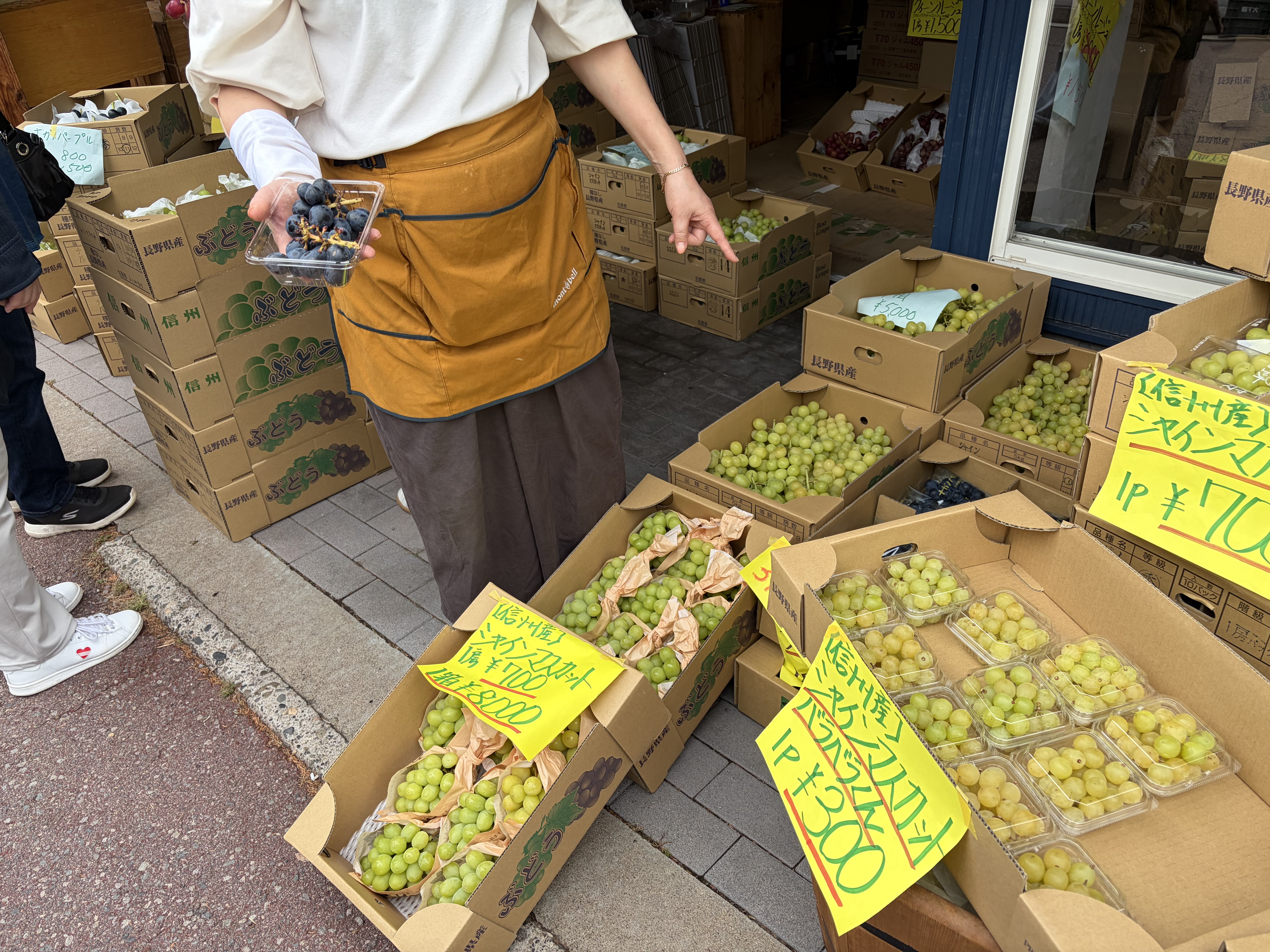店先に箱入りのぶどうが並び、値札を指し示す人物が写っている。