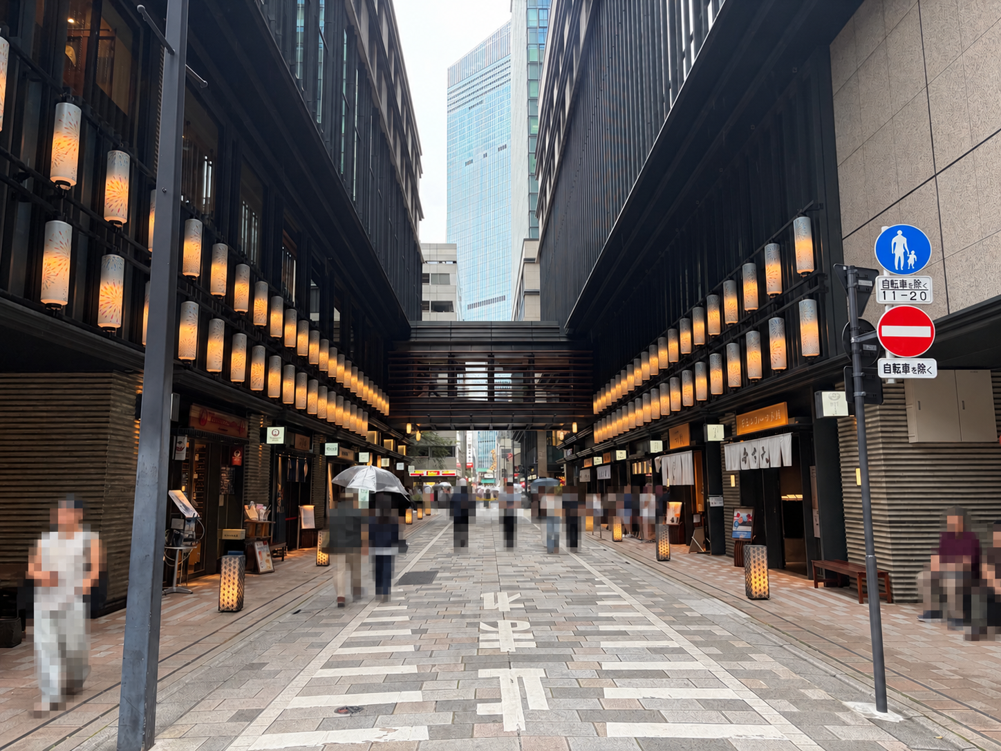 暖色の照明が連なる飲食店街の歩行者通路。高層ビルの間に連絡通路が架かっている。